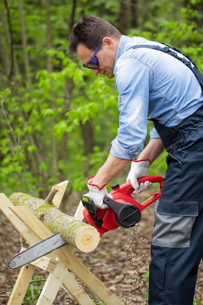 Lumberman in working uniform sawing tree trunk on sawhorse with chainsaw