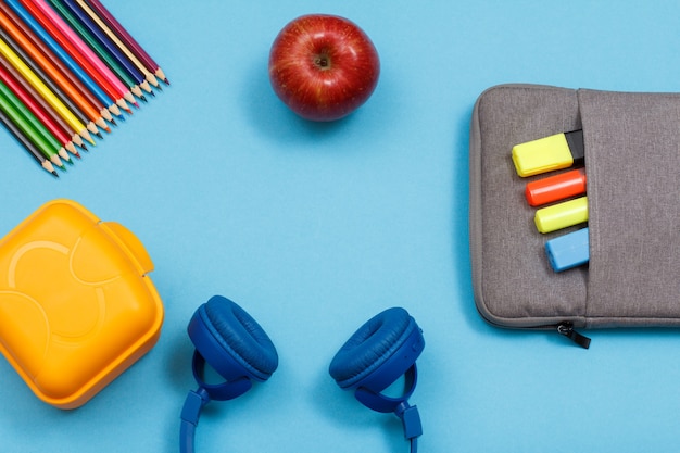 파란색 배경에 색 펠트 펜과 마커가 있는 도시락, 컬러 연필, 사과, 헤드폰, 가방 연필 케이스. 복사 공간이 있는 상위 뷰입니다. 학교 개념으로 돌아가기. 학용품.