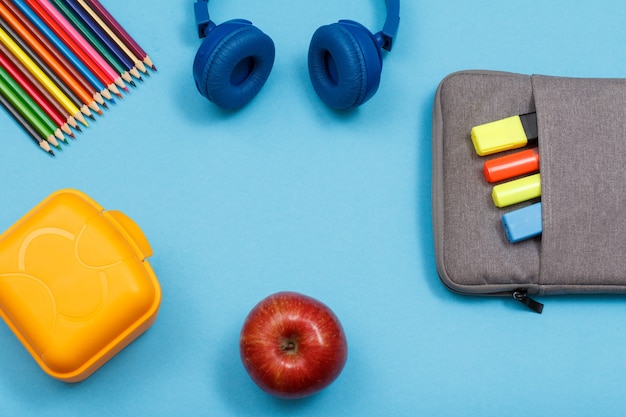 Lunch box, color pencils, headphones, apple and bag-pencil case with color felt pens and marker on blue background. Top view with copy space. Back to school concept. School supplies