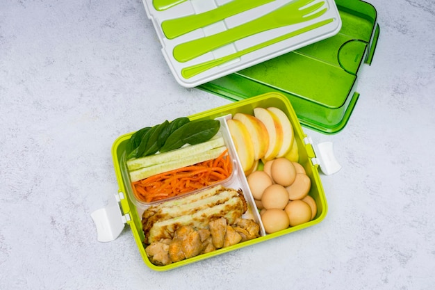 Коробка для завтрака в зеленом цвете со столовыми приборами Закуска с курицей, огурцами, морковью, листьями шпината, нарезанным яблоком и печеньем на десерт, здоровое питание, копия пространства, вид сверху.