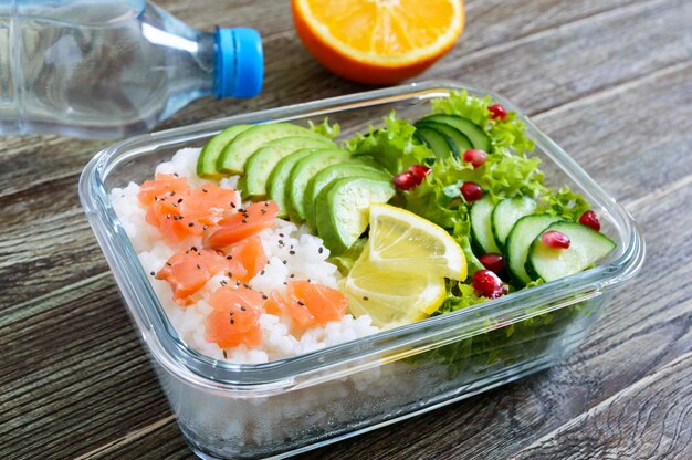 Lunch box: rice, salmon, salad with cucumber, avocado, greens, lemon, fresh orange, and bottle of water on a wooden background. Fitness food. The concept of healthy eating. School lunch box