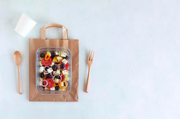 Lunch box with greek salad on the paper bag with wooden fork and spoon, paper cup, on white background, concept of healthy food and zero waste, top view, copy space