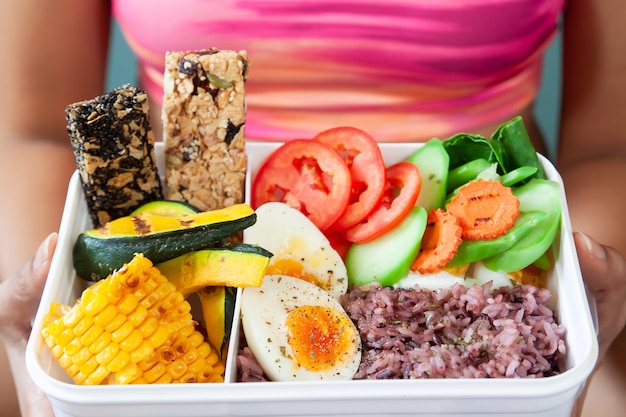 Photo lunch box with rice berry, boiled eggs, vegetables, corn, pumpkin and cereal bars