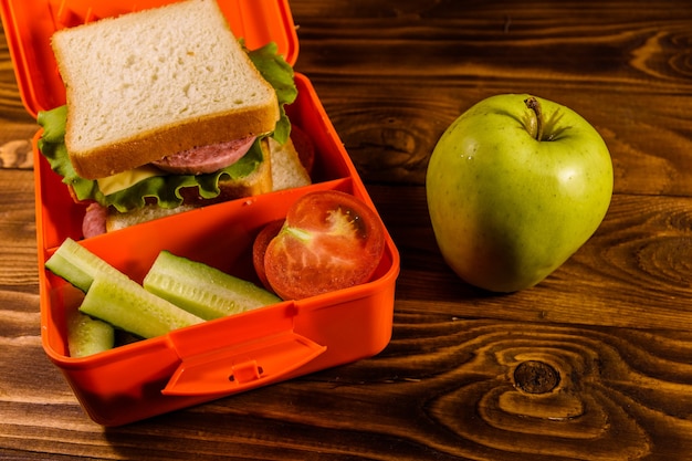 Lunch box with sandwich, cucumbers, green apple and tomatoes on rustic wooden table