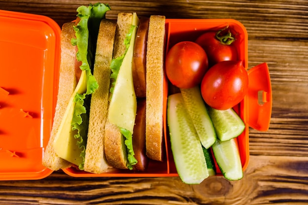 Lunch box with sandwiches cucumbers and tomatoes on rustic wooden table Top view