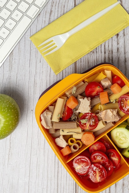 Lunch op de werkplek gezonde pasta met tonijn, kerstomaatjes, wortel, komkommer in lunchbox op werktafel. Thuisvoedsel voor kantoorconcept