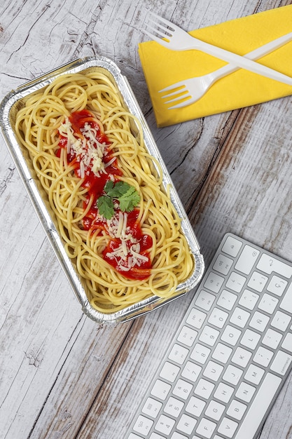Lunch op de werkplek zelfgemaakte en gezonde spaghetti met tomaat en kaas op werktafel met computertoetsenbord. Thuisvoedsel voor kantoorconcept. Afhaalmaaltijden