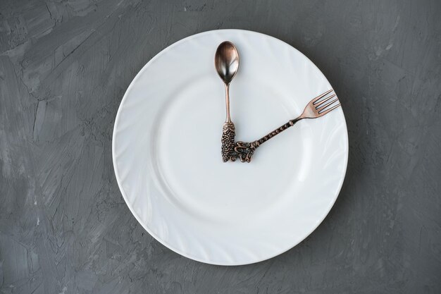 Photo lunch time concept spoon and a fork on a plate in a shape of clock hands time for dinner or breakfast cooking at home or restaurant white plate and clockwise on grey background