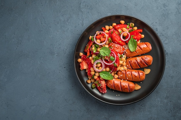 Lunch with vegetable salad with chickpeas and tomatoes and sausages on a dark background