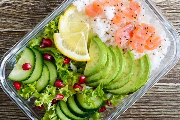 Lunchbox: rijst, zalm, salade met komkommer, avocado, groenen, citroen, chiazaden op een houten achtergrond. fitness eten. het concept van gezond eten. school lunchbox. bovenaanzicht, plat gelegd. detailopname.