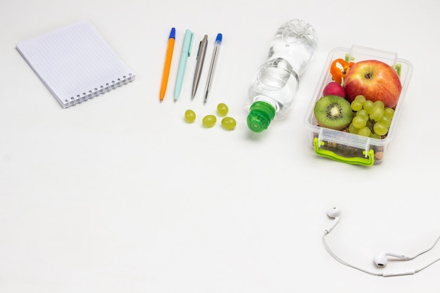 Lunchbox with fruits on table. Notepad Pens, headphones and bottle of water on white
