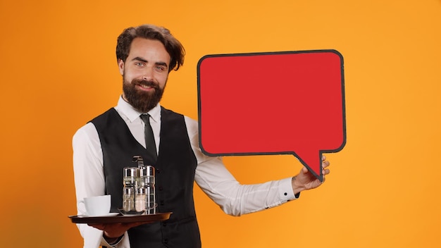 Luxe butler presenteert een lege tekstballon in de studio, werkt in een vijfsterrenrestaurant en houdt een rood marketingadvertentiepictogram vast met een leeg reclamebord. Professionele ober met metalen dienblad voor eten en drinken.