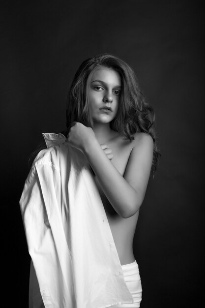 Luxe jong model draagt een wit herenoverhemd en poseert met blote schouders in de studio. Monochrome opname