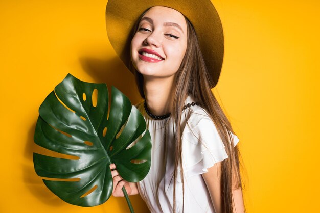 Luxe jong vrouwelijk model met lang haar houdt een groen blad, hoed op haar hoofd, poserend op een gele achtergrond