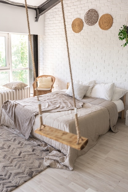 Luxe slaapkamerontwerp in een rustiek huisje in een minimalistische stijl. witte muren, panoramische ramen, houten decoratie-elementen aan het plafond, touwschommels in het midden van een ruime kamer.