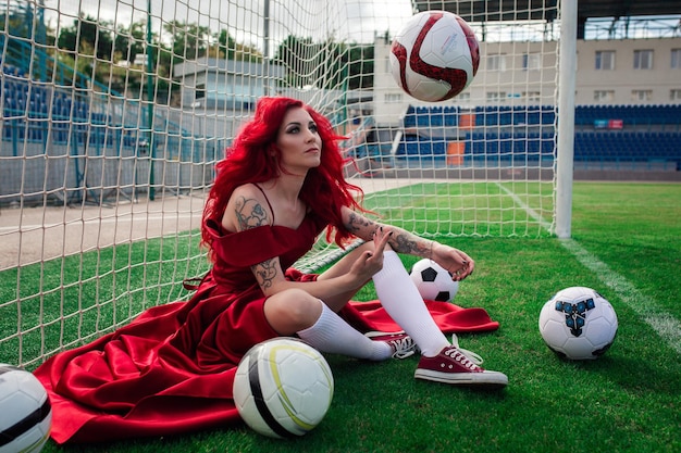 Luxe vrouw met rood haar en in een rode jurk speelt op het voetbalveld Idee en concept van een combinatie van sport en schoonheid ongebruikelijke presentatie