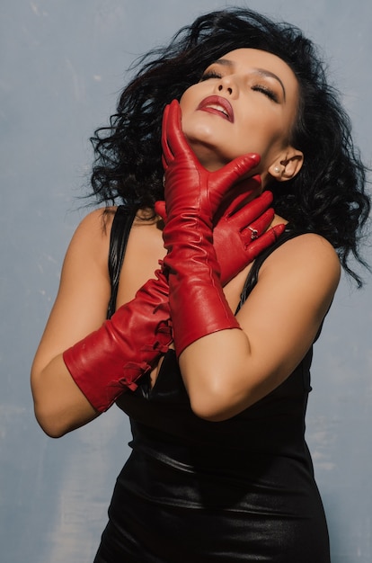 Роскошная азиатская женщина в черном кожаном платье и красных перчатках держится за горло. Доминирующая фетиш-леди.