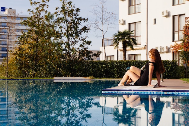 A luxurious attractive young woman in a black dress enjoys relaxing by the blue pool in the sun