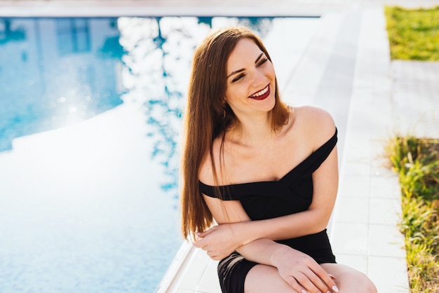 Luxurious attractive young woman in a black dress sitting by the pool, laughing, rich life