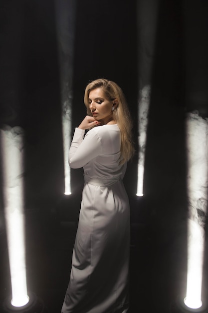 Luxurious blonde plus size with white long hair posing in a long white dress on a dark stage in the smoke with the light.