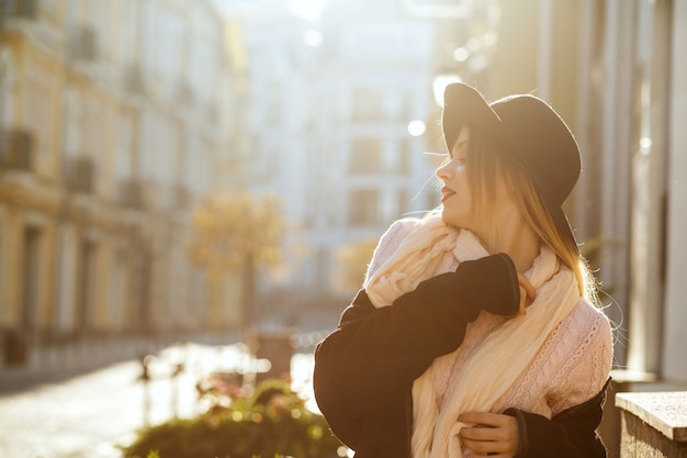 Luxurious blonde woman wearing sweater and hat posing at the old street. Space for text