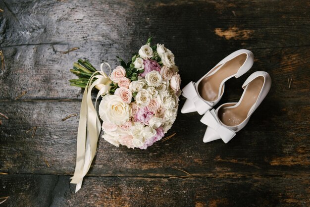 A luxurious bridal bouquet with white roses next to shoes and wedding invitations on a wooden background