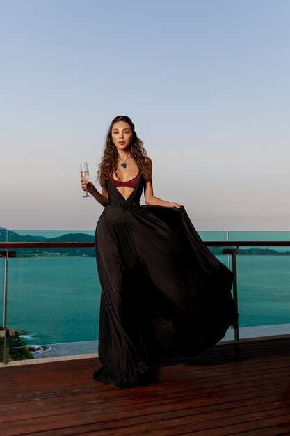 고급스러운 갈색 발코니에 와인 한 잔과 비싼 보석에 검은 긴 드레스를 입고 포즈. 열대 섬과 바다의 고급스러운 전망