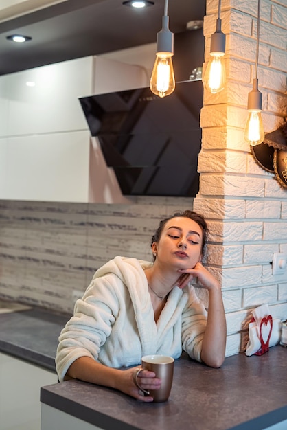 Luxurious lady in a snowwhite robe holds a cup of coffee to cheer herself up