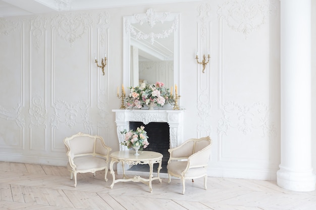 Luxurious light interior in the Baroque style. A spacious room with a road chic beautiful furniture, a fireplace and flowers. plant stucco on the walls and light wood parquet