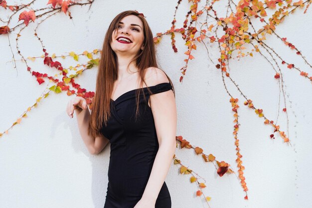 Luxurious long-haired woman in a long black dress laughing, posing against the wall in her garden