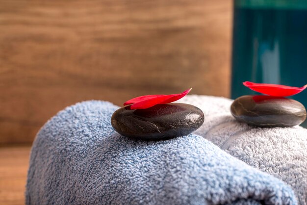 Photo luxurious spa arrangement for two with a a blue and white folded soft towel, black massage stones with red rose petal on top and a bottle of massage oil or moisturizing lotion.