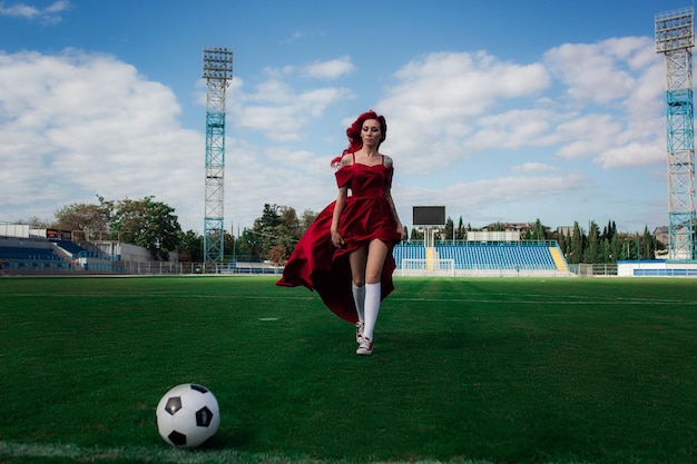 Luxurious woman with red hair and in a red dress plays on the football field Idea and concept of a combination of sports and beauty unusual presentation