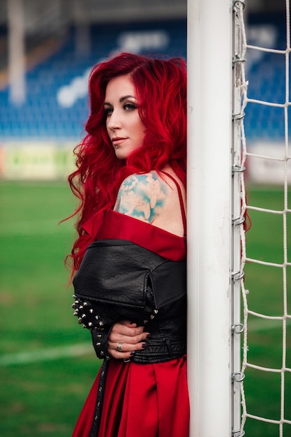Luxurious woman with red hair and in a red dress plays on the football field Idea and concept of a combination of sports and beauty unusual presentation