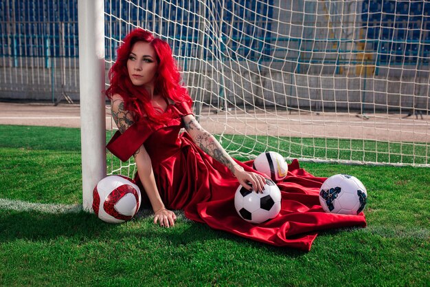 Luxurious woman with red hair and in a red dress plays on the football field Idea and concept of a combination of sports and beauty unusual presentation