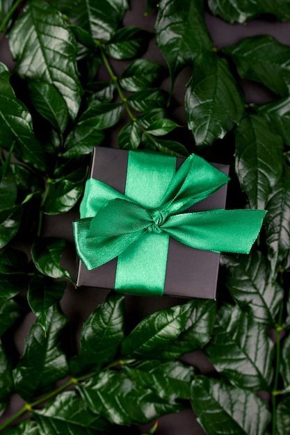 Luxury black gift box with green ribbon on a dark background with leaves on the sides, flat lay, nature concept.