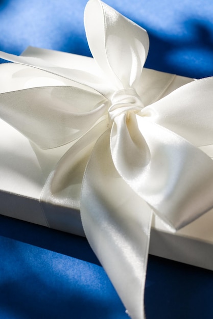 Luxury holiday white gift box with silk ribbon and bow on blue background luxe wedding or birthday present