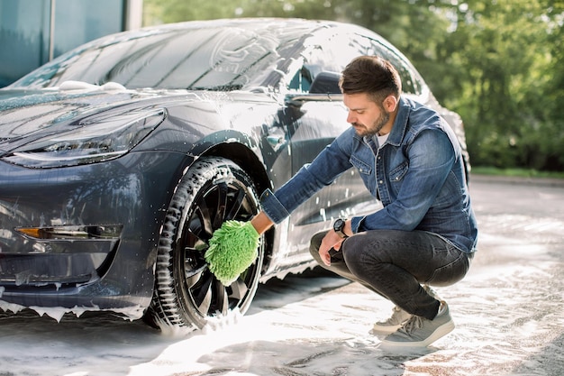Luxury modern electric car in soap foam outdoors at car wash service Side view of handsome young Caucasian man using green microfiber car wash mitt for cleaning rims outdoors
