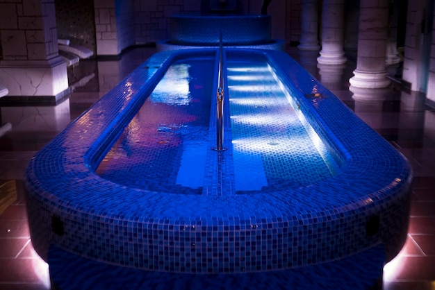 A luxury pillared spa hall with the illuminated plunge pool in the centre An empty beautiful bluetiled spa pool of the deluxe hotel Wall columns sauna design Illuminated swimming pool in spa