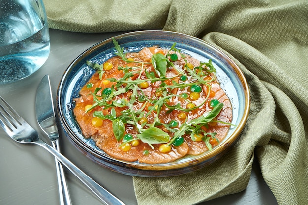 Luxury salad of slightly salted crudo salmon with arugula and sauces in a blue plate on a wooden table. Antipasti seafood