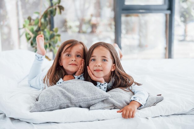 Photo lying down on bed and looking at the camera two cute little girls indoors at home together children having fun