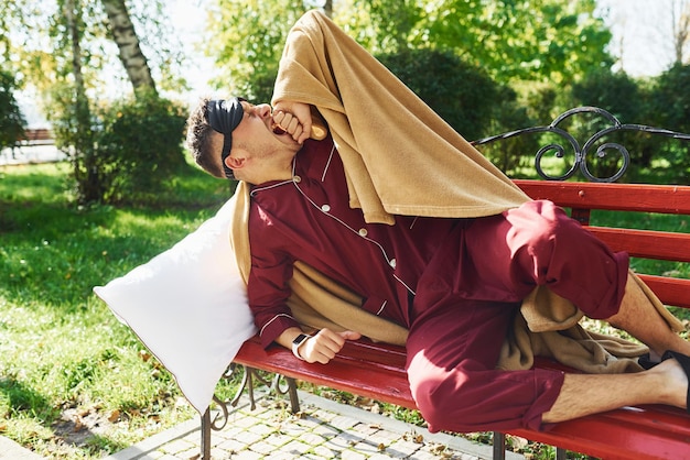 Lying down on bench in park Young man in pajama is outdoors on the street Feels sleepy