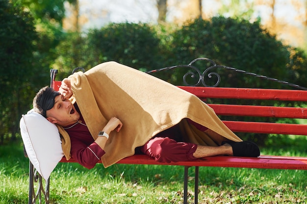 Lying down on bench in park Young man in pajama is outdoors on the street Feels sleepy