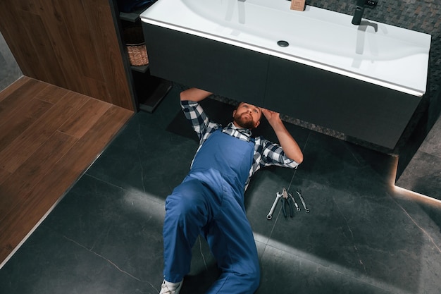 Photo lying down on the floor fixing the siphon plumber in blue uniform is at work in the bathroom