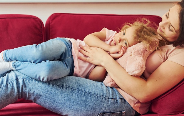 写真 赤いソファに横になっている若い母親と彼女の小さな娘は、自宅で一緒にカジュアルな服を着ています