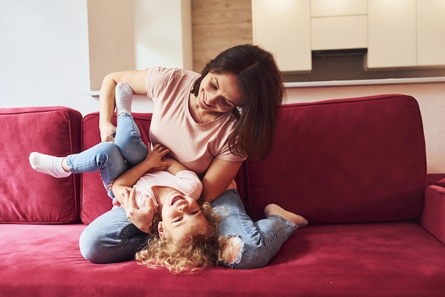 赤いソファに横になっている若い母親と彼女の小さな娘は、自宅で一緒にカジュアルな服を着ています