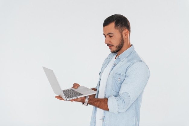 Maakt gebruik van laptop Jonge knappe man die binnenshuis tegen een witte achtergrond staat