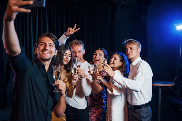 Maakt selfie. Groep vrolijke vrienden die nieuwjaar binnenshuis vieren met drankjes in handen.