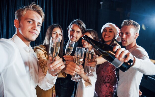 Maakt selfie. Groep vrolijke vrienden die nieuwjaar binnenshuis vieren met drankjes in handen.