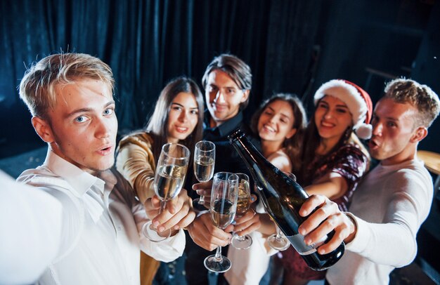 Maakt selfie. Groep vrolijke vrienden die nieuwjaar binnenshuis vieren met drankjes in handen.