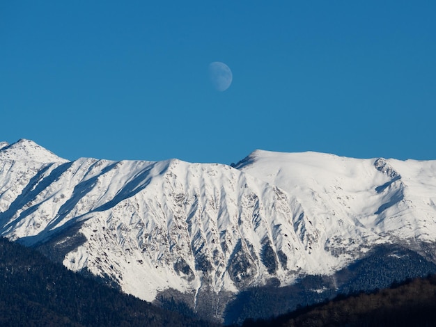 Maan op heldere blauwe lucht boven besneeuwde bergen op zonnige dag